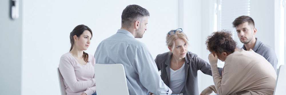 Therapist with glasses on head comforting a member of a bipolar support group as the other members look on